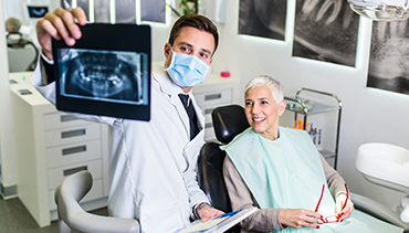 Handsome male dentist checking x-ray image or scan while beautiful senior woman receiving a dental treatment.